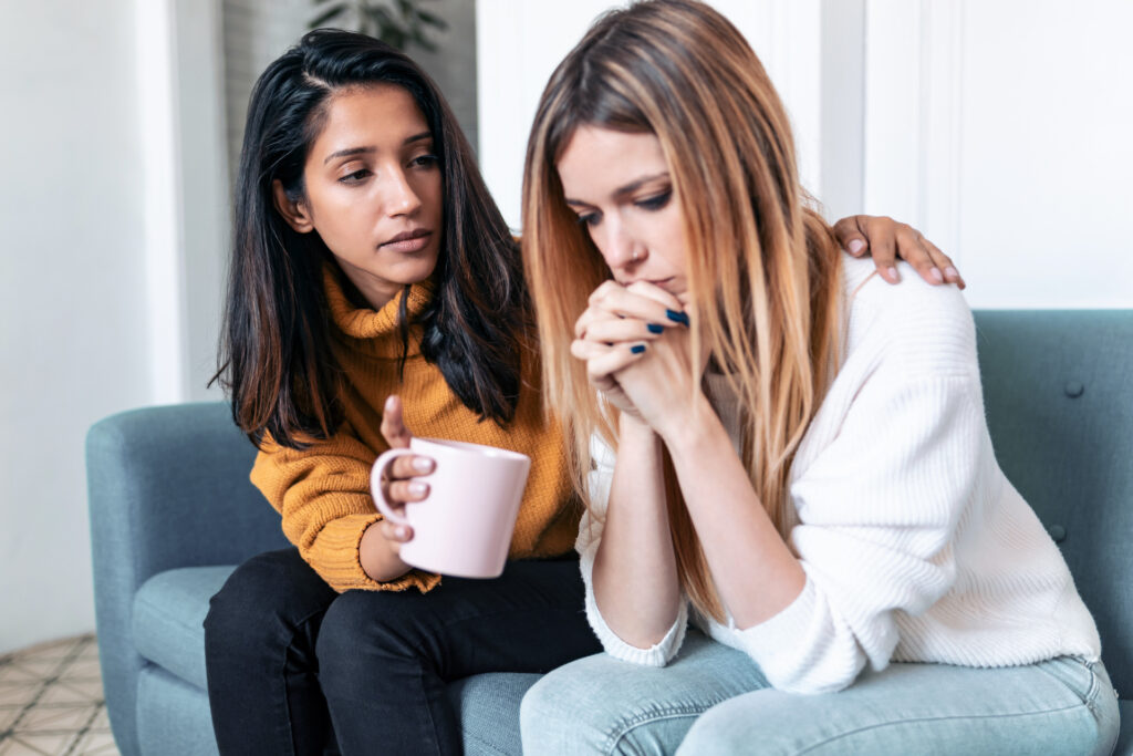 2 young woman sitting on a couch with concerned expressions on their faces. One woman offering support to the other.